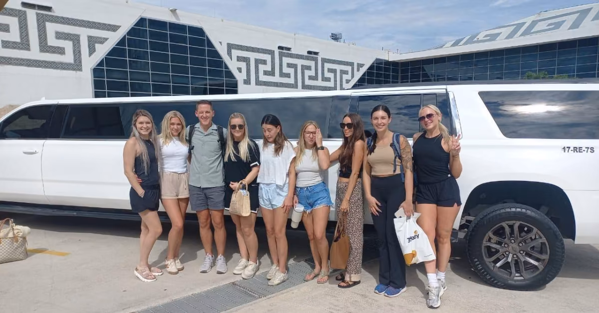 Eight people, seven women and one man, smile in front of a white limo parked outside a modern building with geometric designs.