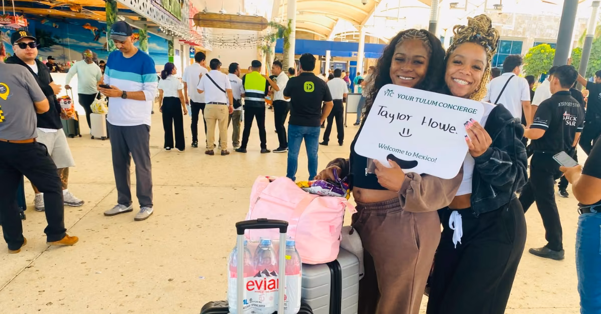 Two women at an airport smile and hold a welcome sign by their luggage, with others waiting in the background.