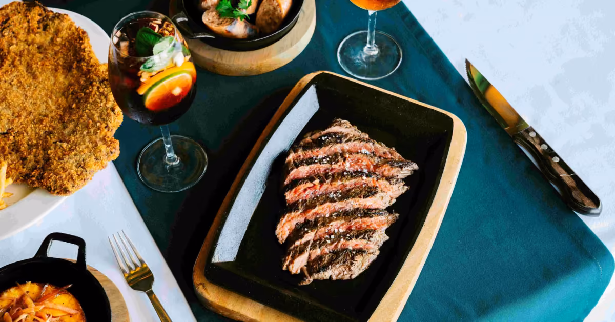 Table with sliced steak, fried bites, tapas, and two glasses of fruit sangria on a green and white tablecloth.