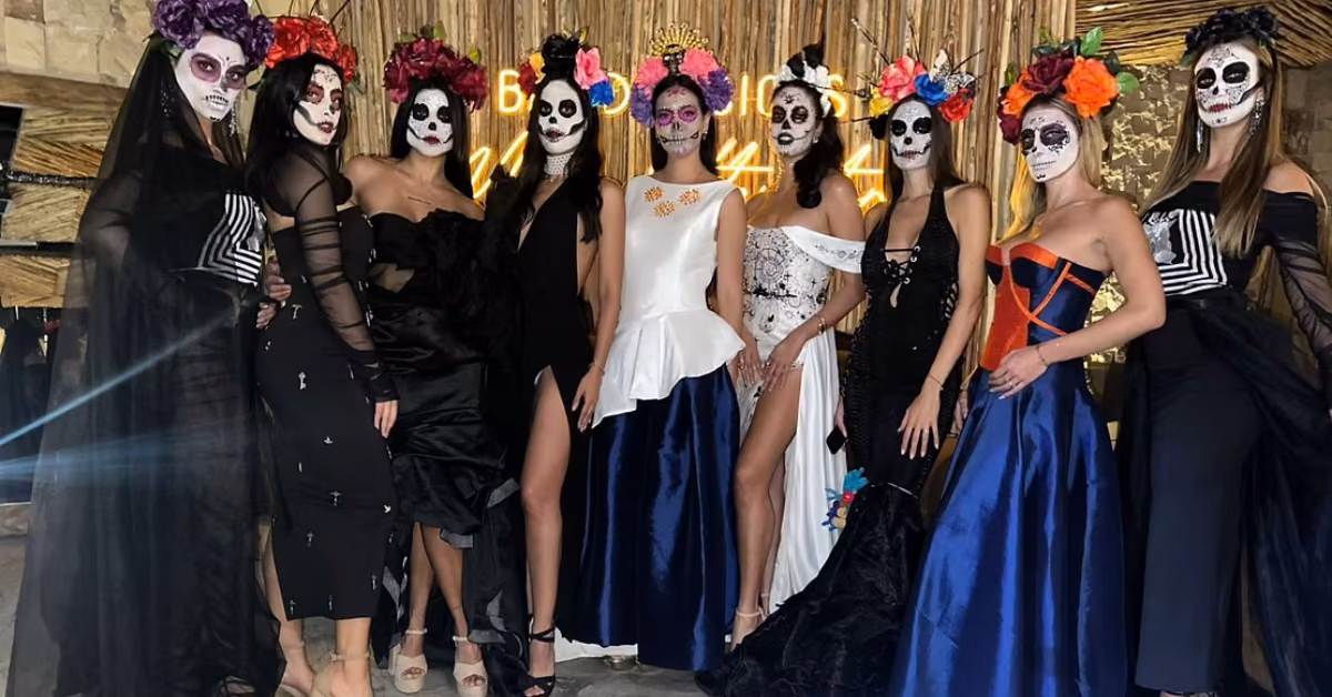 Eight women in elegant dresses and Day of the Dead skull makeup pose before a glowing, festive backdrop at Chambao Restaurant.