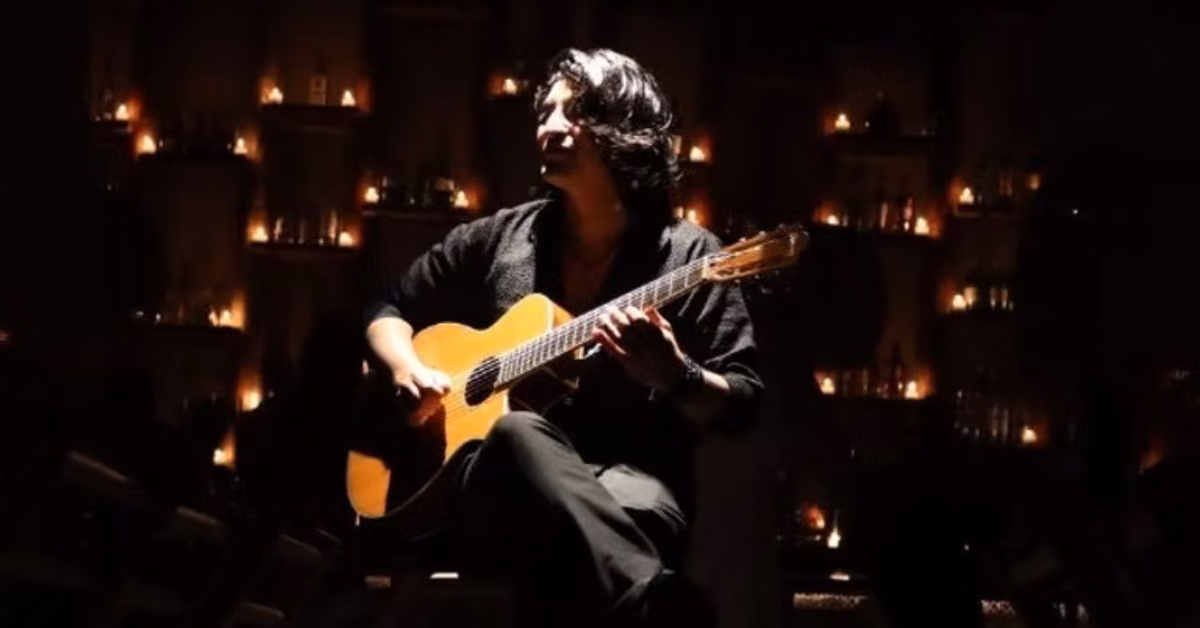 A person with long dark hair plays acoustic guitar in a dim room, surrounded by glowing candles for an intimate, cozy feel.