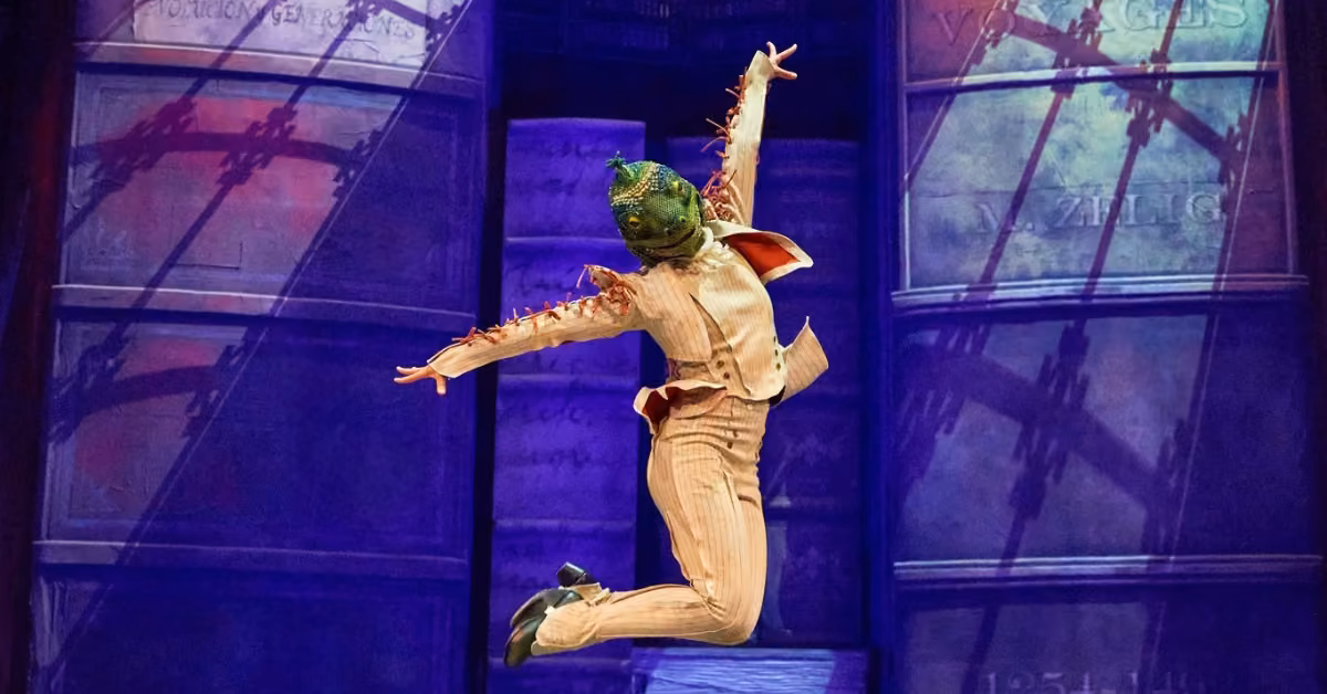 A performer in a tan pinstripe suit and green lizard mask leaps mid-air on stage, books and purple lights behind them.