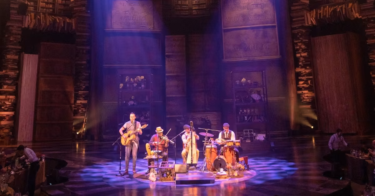 Four musicians perform on stage under purple lights, surrounded by tall bookshelves in a dramatic, library-themed setting.