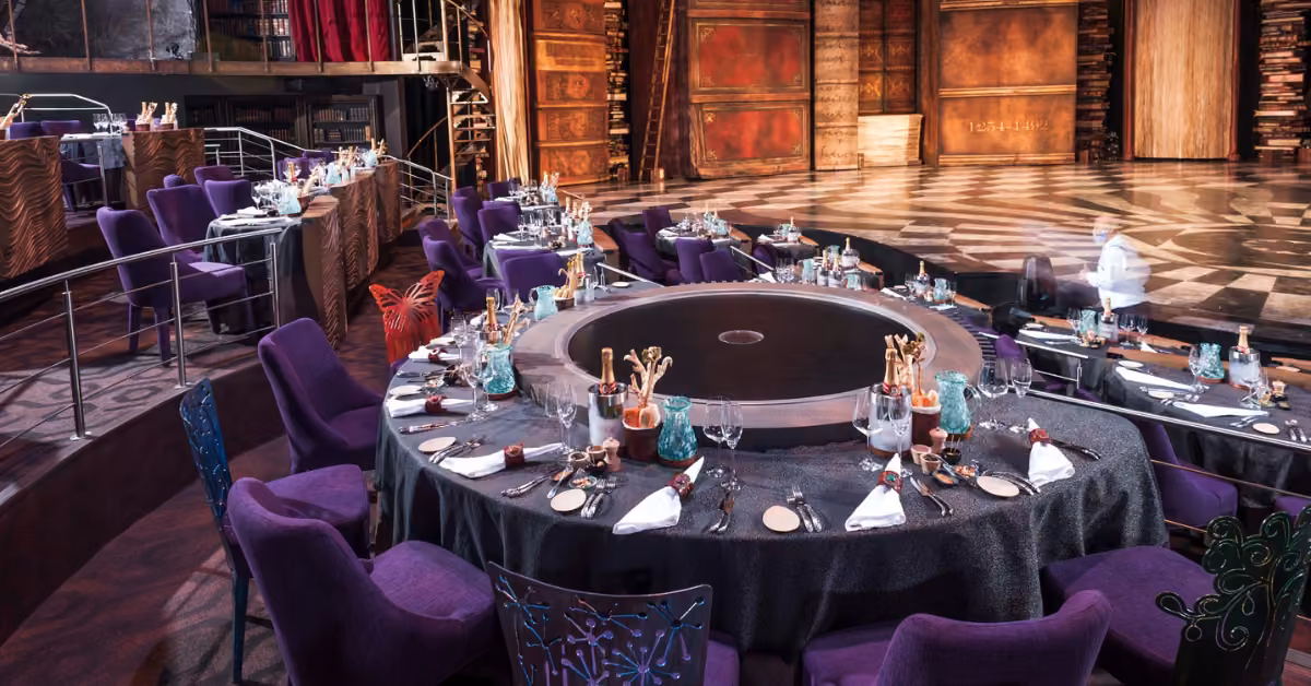 Round tables with elegant settings and purple chairs encircle a stage in a dim, lavish theater set for an upscale dinner show.