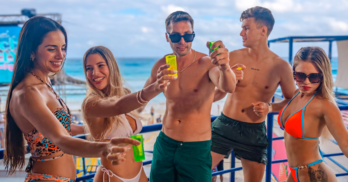 Five friends in swimsuits cheer with colorful drinks, smiling at a beach club with the ocean and blue sky behind them.