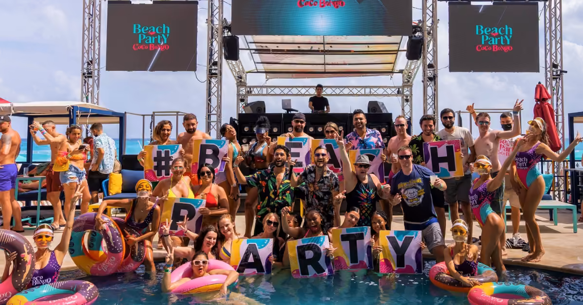 People in swimsuits pose in and around a pool, holding “BEACH PARTY” signs at Coco Bongo Beach Club, DJs and banners behind.