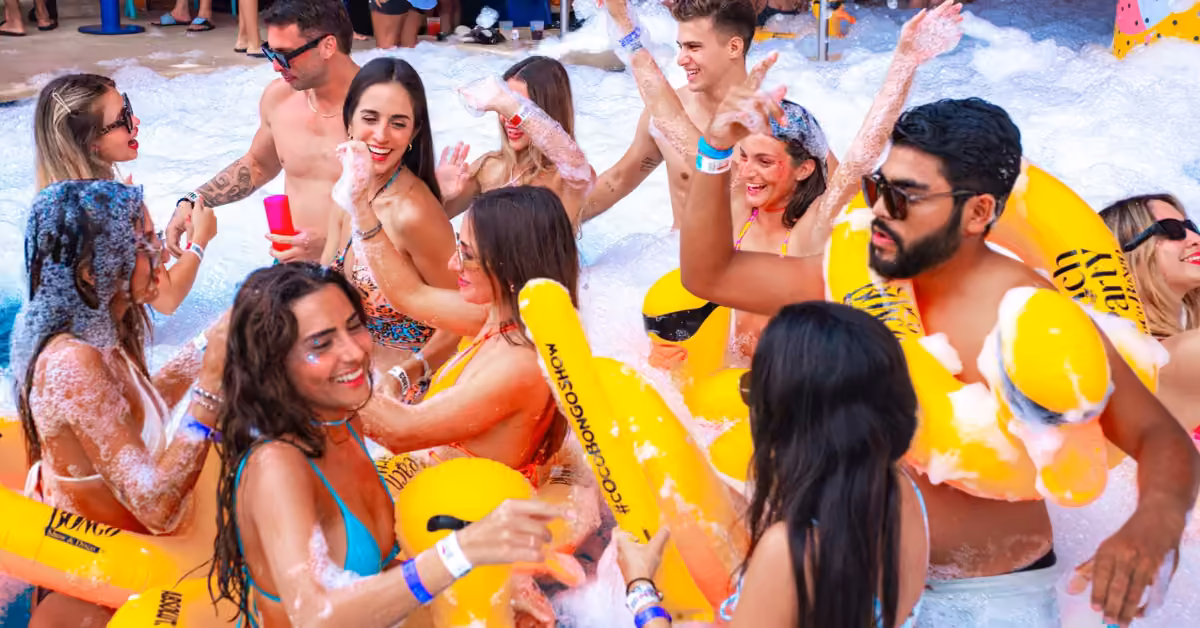Young adults in swimwear laugh and dance at a lively foam party, surrounded by bubbles and yellow inflatables outdoors.