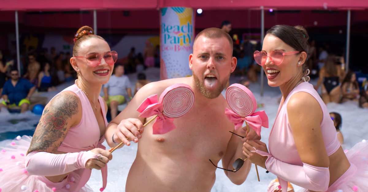 Two women in pink and heart-shaped glasses stand beside a shirtless man with pink lollipops at a lively pool party.