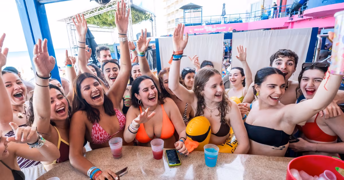 Young people in swimsuits laugh and raise their hands, holding colorful drinks at a lively poolside bar party.