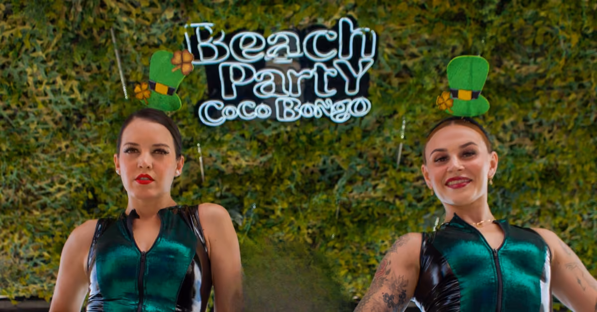 Two women in shiny green outfits with clover headbands pose by a leafy backdrop and “Beach Party Coco Bongo” sign.