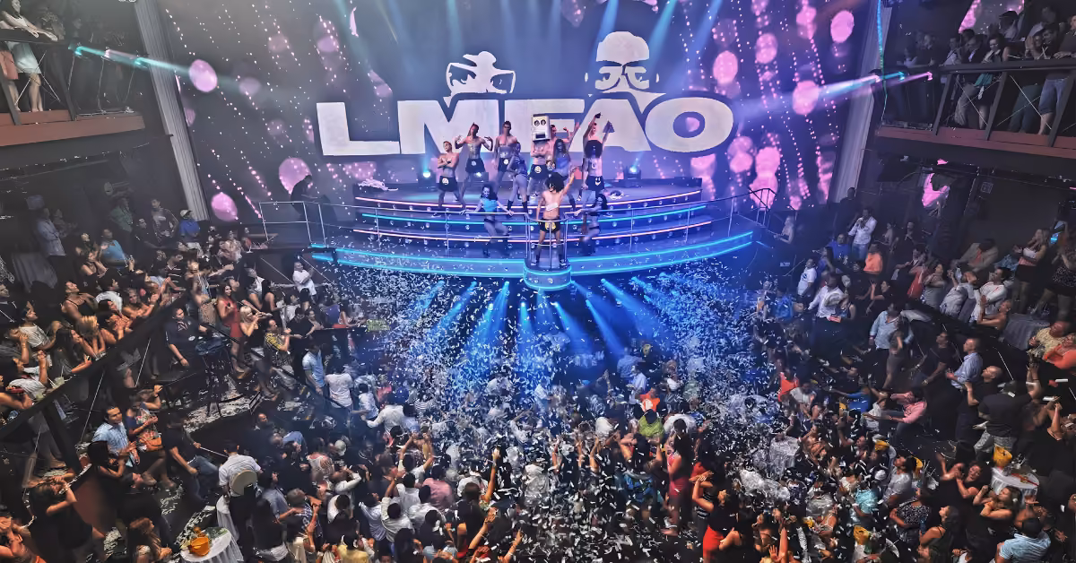 Crowds dance amid confetti in a vibrant night club as performers light up the stage and LMFAO shines in the backdrop.