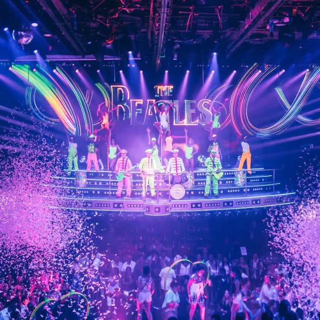 Performers in colorful costumes dance under a neon Beatles sign as confetti falls on a cheering crowd lit by purple lights.