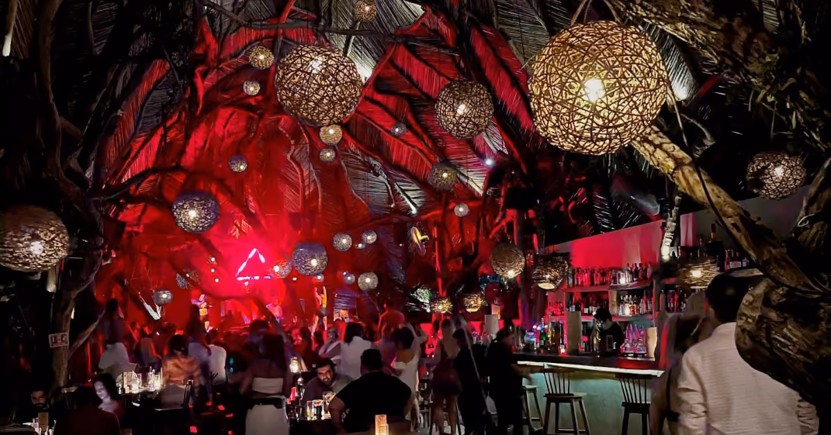 A lively, dim bar with a thatched ceiling, glowing lanterns, people socializing, and red stage lights in the background.