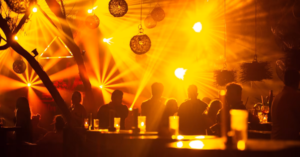 People gather at tables in the dimly lit Confessions Night Club, with warm stage lights and hanging lamps overhead. Glasses on tables.