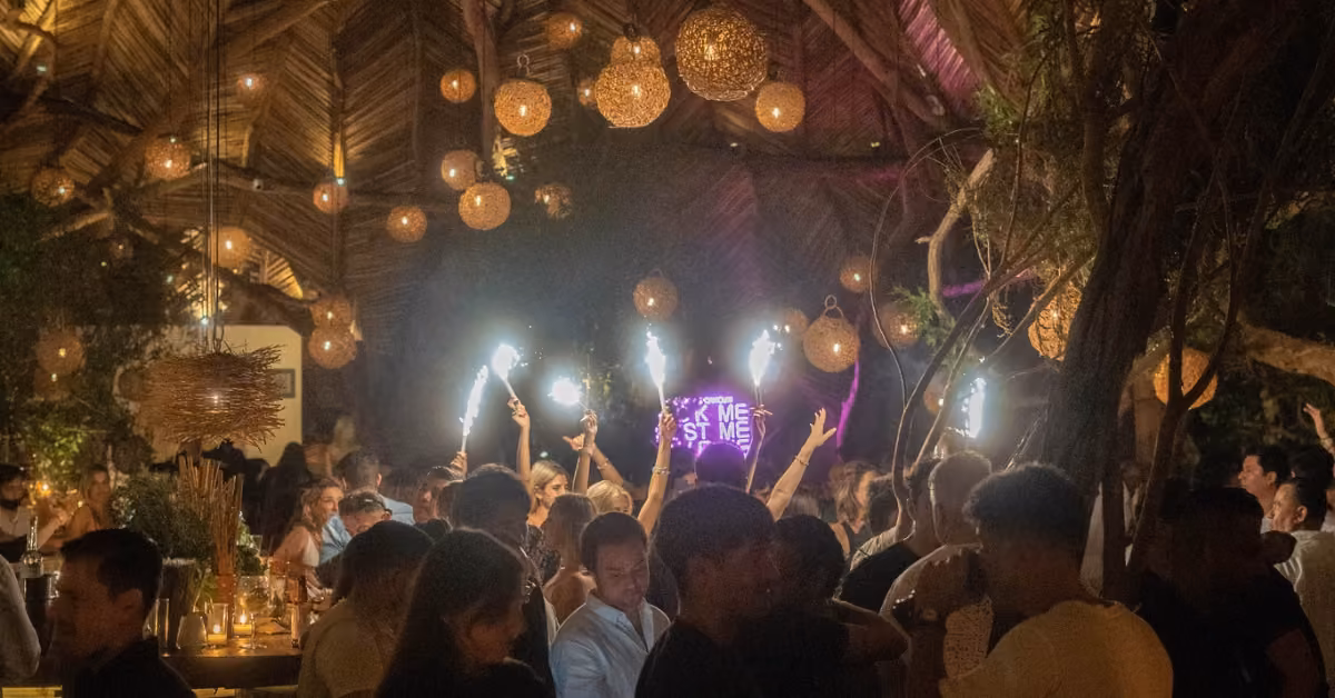 People mingle at a party under warm lights, holding sparklers, with a pink neon sign and tropical wooden decor in the background.