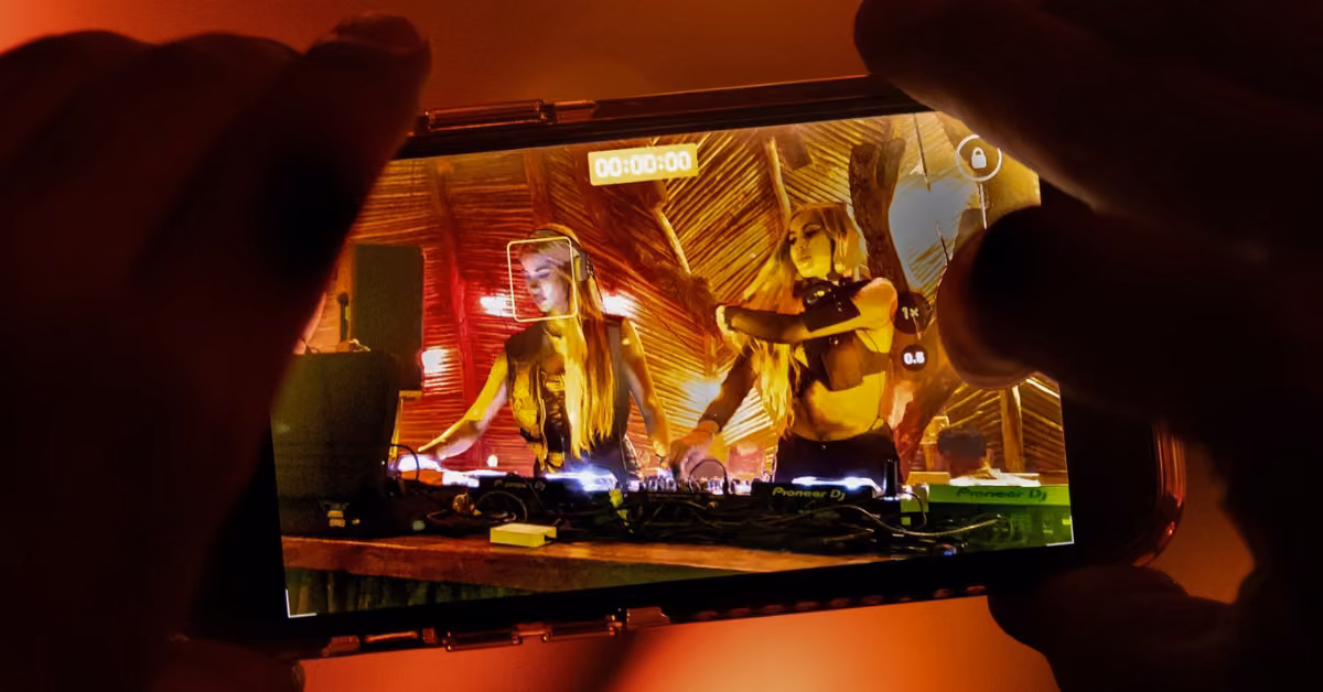 A phone screen captures two DJs performing at a warmly lit club with palm leaves, shown through the camera interface.