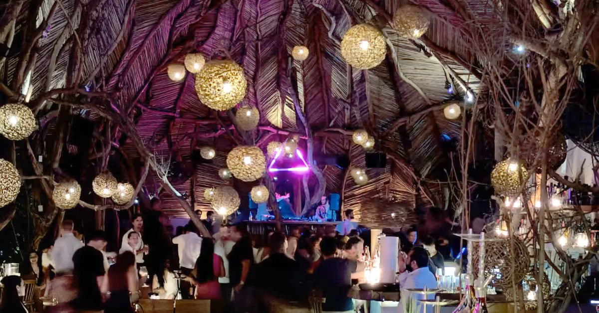 A vibrant bar with a thatched ceiling, glowing lanterns, purple lighting, and people socializing in a festive atmosphere.
