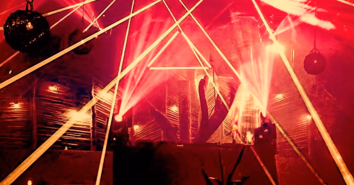 A night club with red lighting and beams crossing above a DJ booth, creating drama. Hanging lights and silhouettes fill the background.