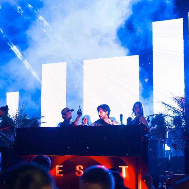 A DJ performs at a booth with vivid LED screens, colorful lights, and smoke as people dance in a lively night club atmosphere.
