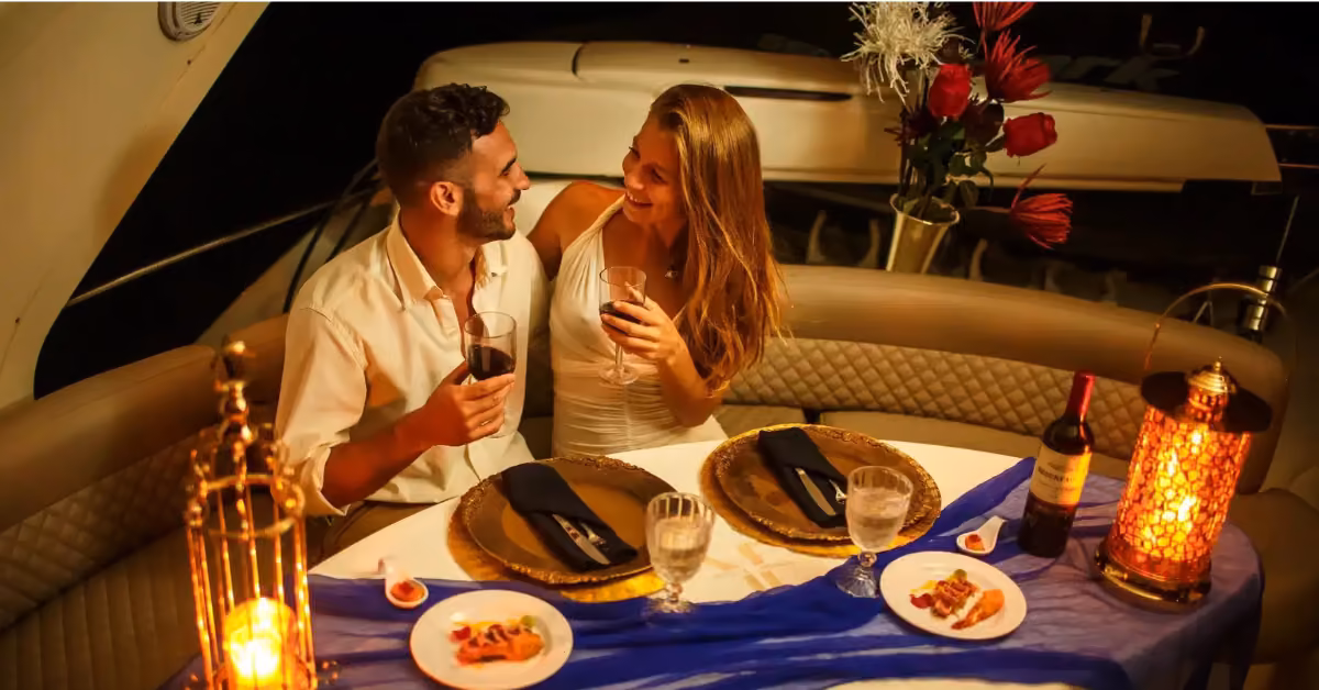 A smiling couple sits together on a yacht, toasting with red wine at a candlelit dinner table adorned with flowers and food.