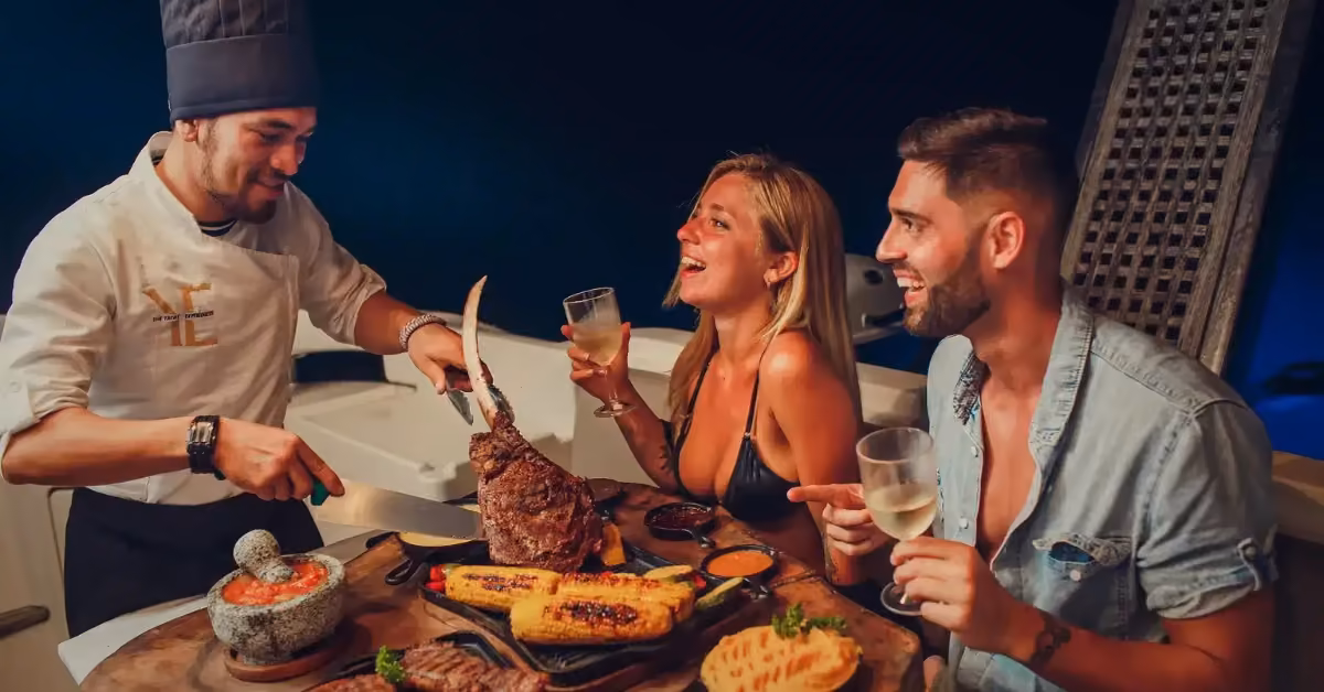 A chef in a tall hat slices steak tableside for a smiling couple with white wine on a dinner cruise, corn and bread on the table.