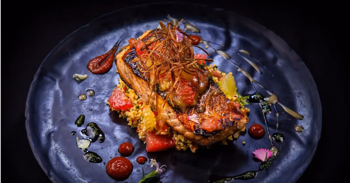 Grilled fish with crispy strips on couscous, grapefruit slices, and garnished with sauces, herbs, and edible flowers.