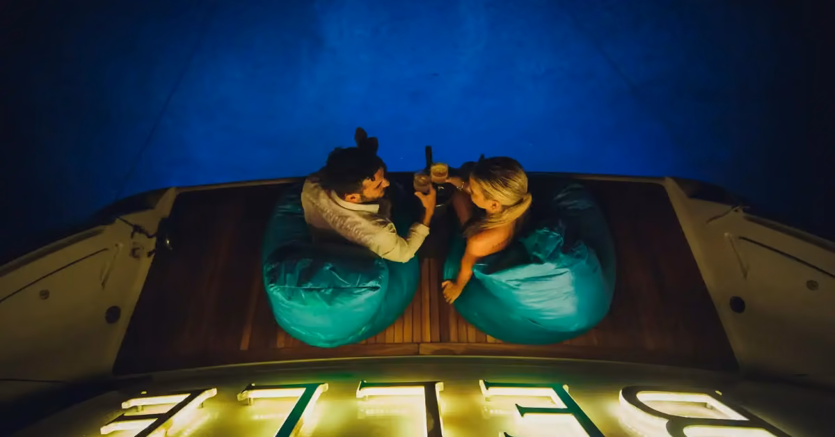 A man and woman relax on turquoise bean bags at night on a boat, drinks in hand, with warm lights and a glowing BELLE sign.