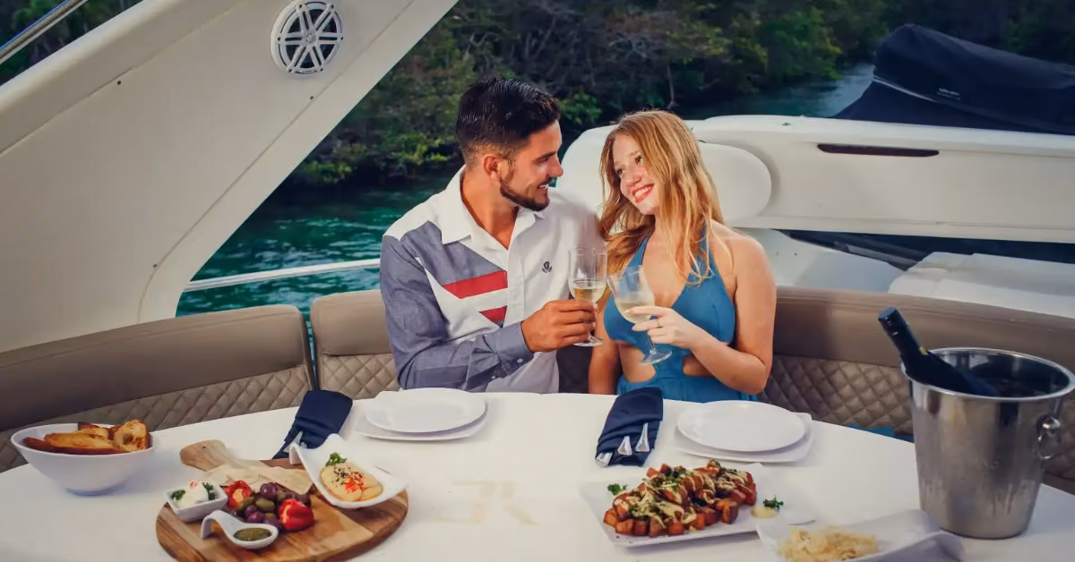 A smiling man and woman toast wine on a yacht, enjoying gourmet food with water and green scenery in the background.