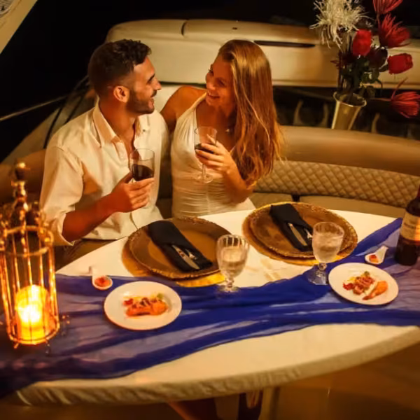 A couple sits close, smiling and sharing wine and appetizers at a candlelit table with flowers and soft romantic lighting.