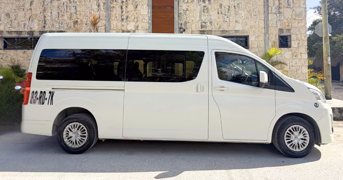 A white van with tinted windows and code 33-RD-7K is parked by a stone building with a wooden door and small plants nearby.