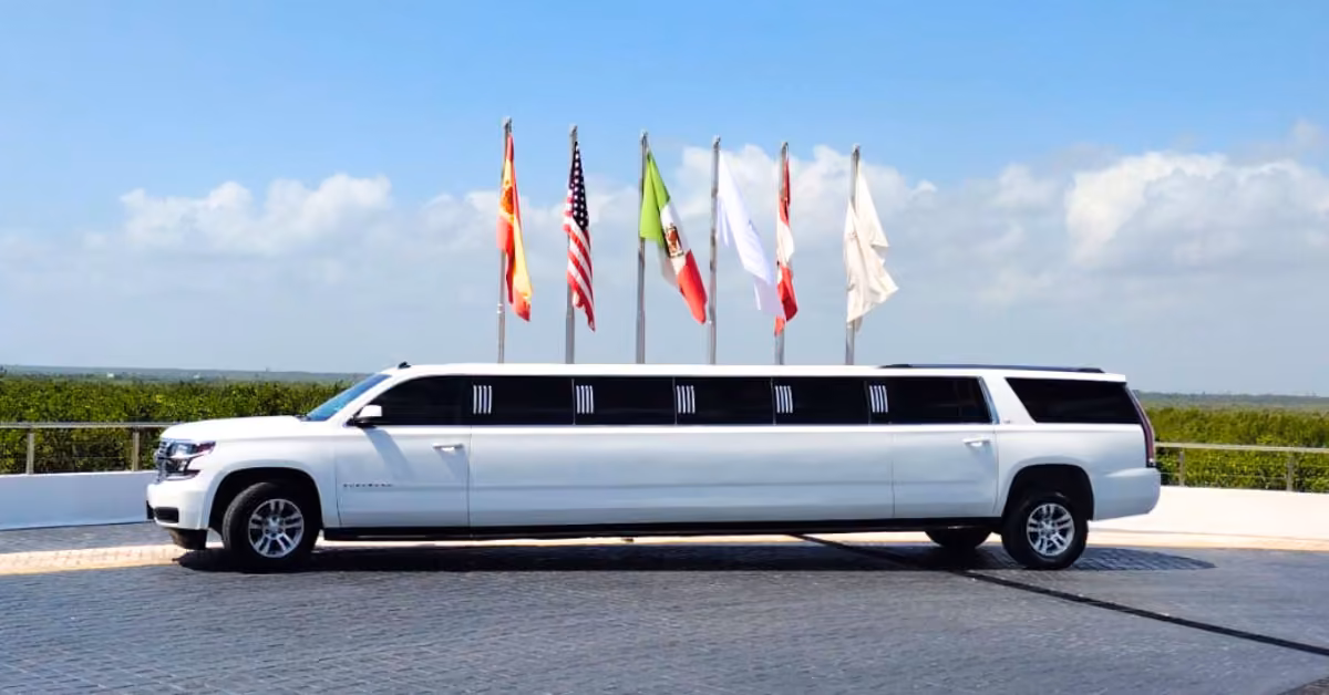 A white stretch SUV limousine is parked on pavement with several flags, including the American flag, under a partly cloudy sky.