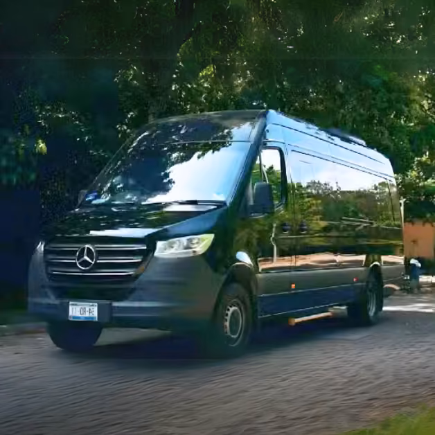 A black Mercedes-Benz Sprinter van drives along a paved road lined with trees and greenery, sunlight glinting on its windows.