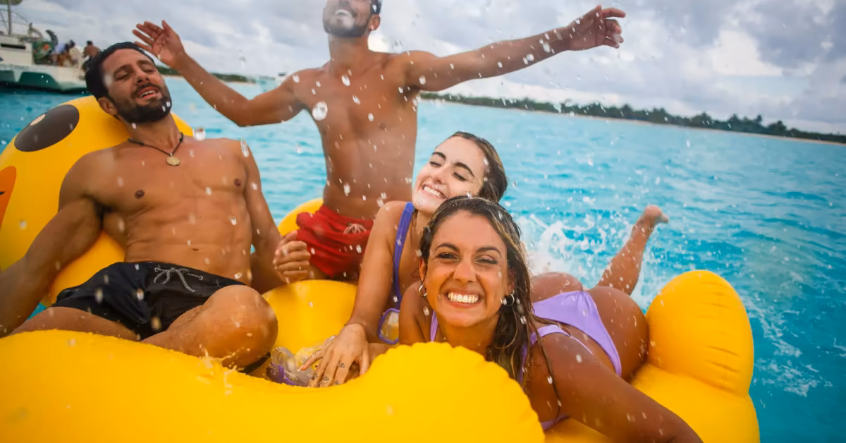 Four young adults laugh and splash on a large yellow float in bright blue water, enjoying a sunny, carefree day together.