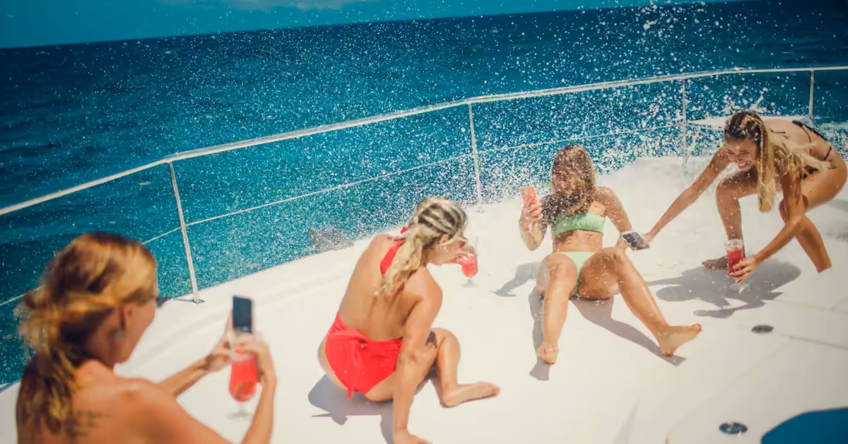 Four women in swimsuits laugh and splash water, holding drinks and enjoying a sunny day together on a cruise.