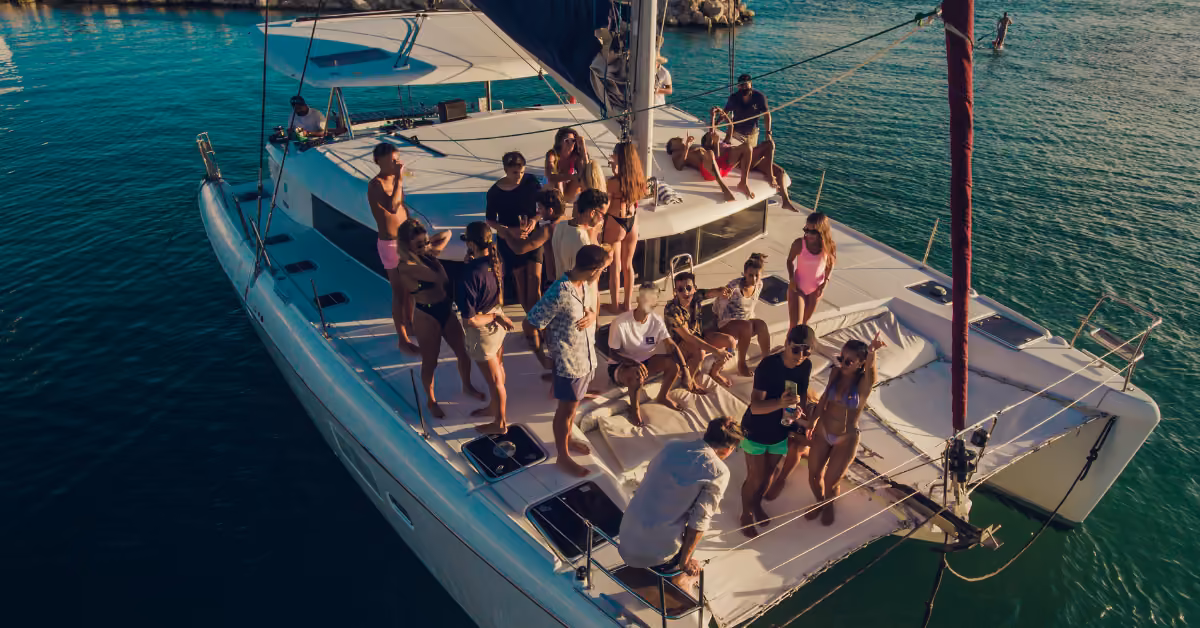 People in swimsuits and summer clothes relax and chat on a white catamaran, anchored in clear water on a sunny day.