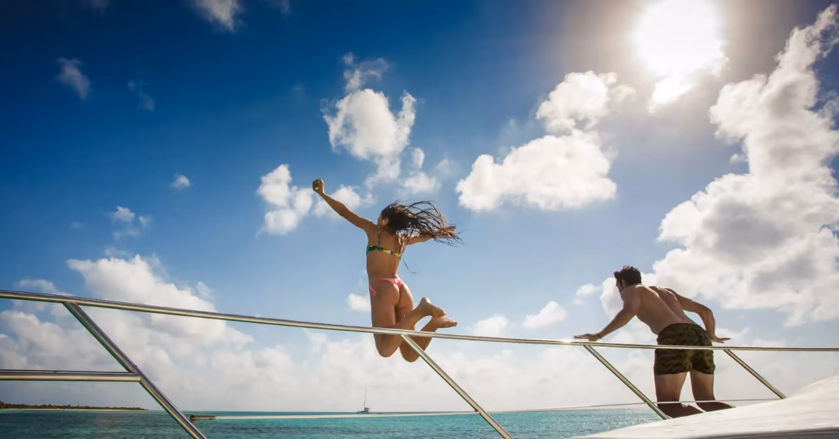 A woman in a bikini leaps off a boat into turquoise water as a man watches, with the sun shining in a partly cloudy sky.