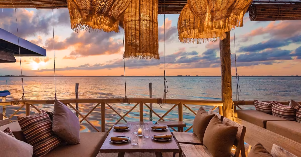 Outdoor dining at sunset with cushioned sofas, set table for four, woven lights overhead, and sea view reflecting warm light.