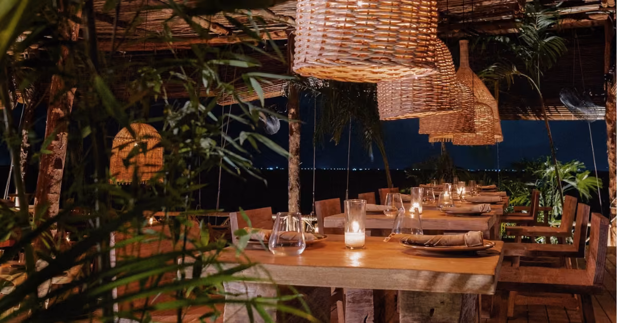 A cozy outdoor restaurant at night with wooden tables, wicker lights, candles, and lush plants creating a warm, inviting vibe.