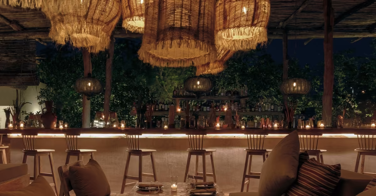 A cozy bar at night with wooden stools, warm lighting, pendant lamps, cushioned seats, and trees outside; candles on the counter.