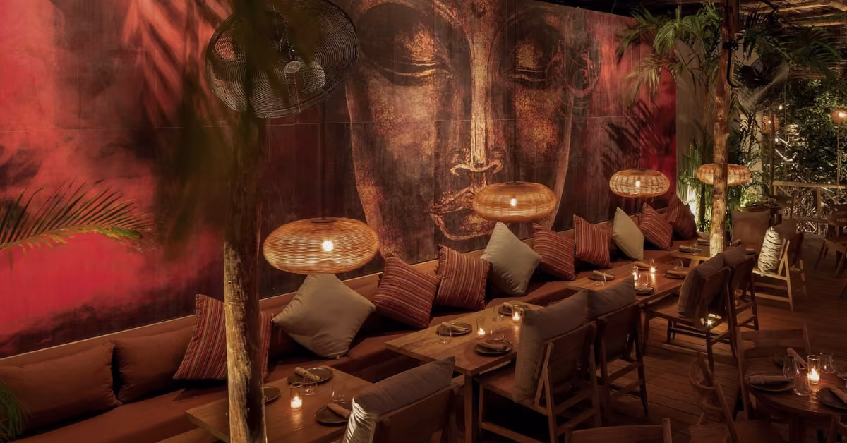 A warm, stylish restaurant with wood tables, brown cushions, woven pendant lights, lush plants, and a serene Buddha mural.
