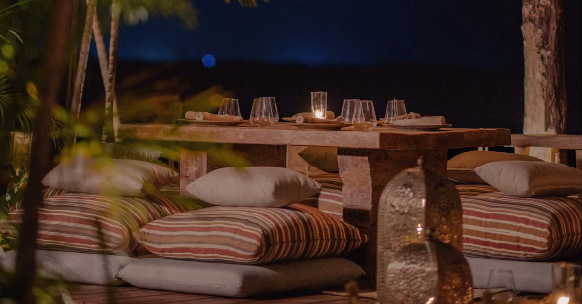 A rustic wooden table with dinnerware, candles, and striped floor cushions creates a cozy, eclectic outdoor dining scene at night.