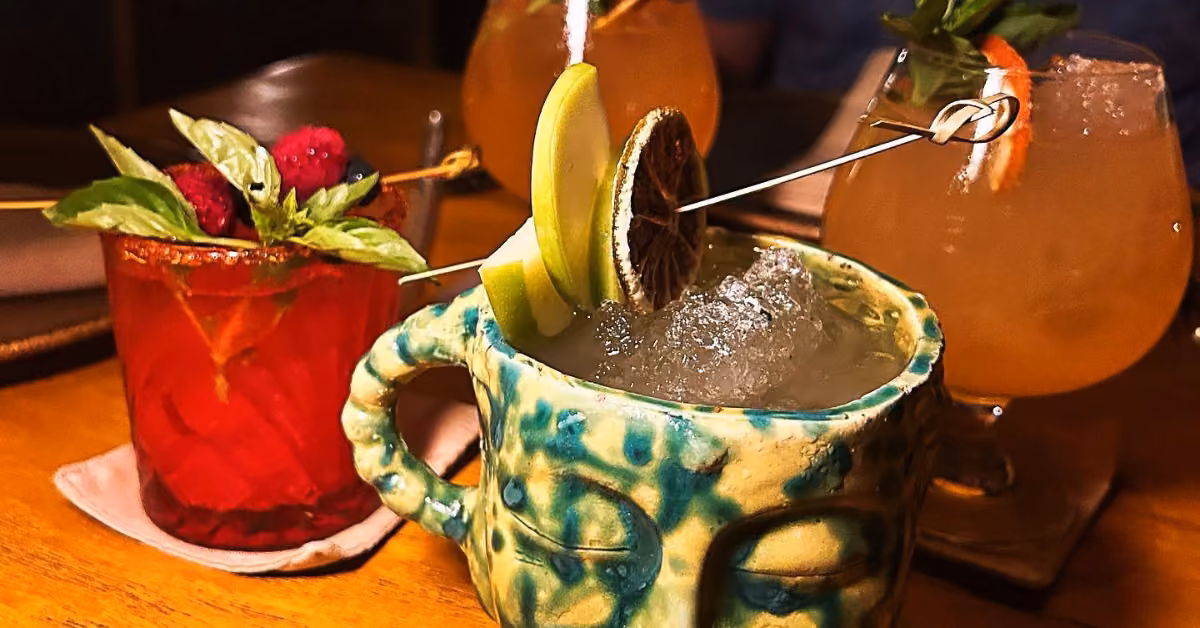 Three cocktails: a red drink with berries and mint, a green drink in a ceramic mug, and a pale orange citrus-topped drink.