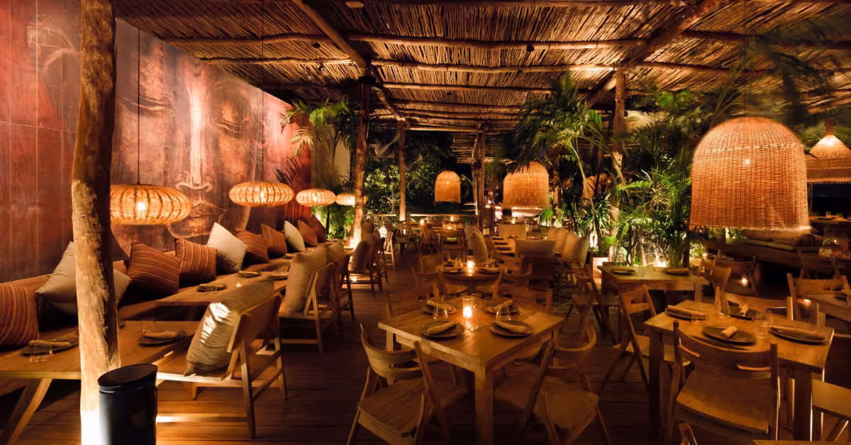 A cozy, welcoming restaurant with wooden tables, wicker lamps, tropical plants, plush seating, and artistic mural decor.