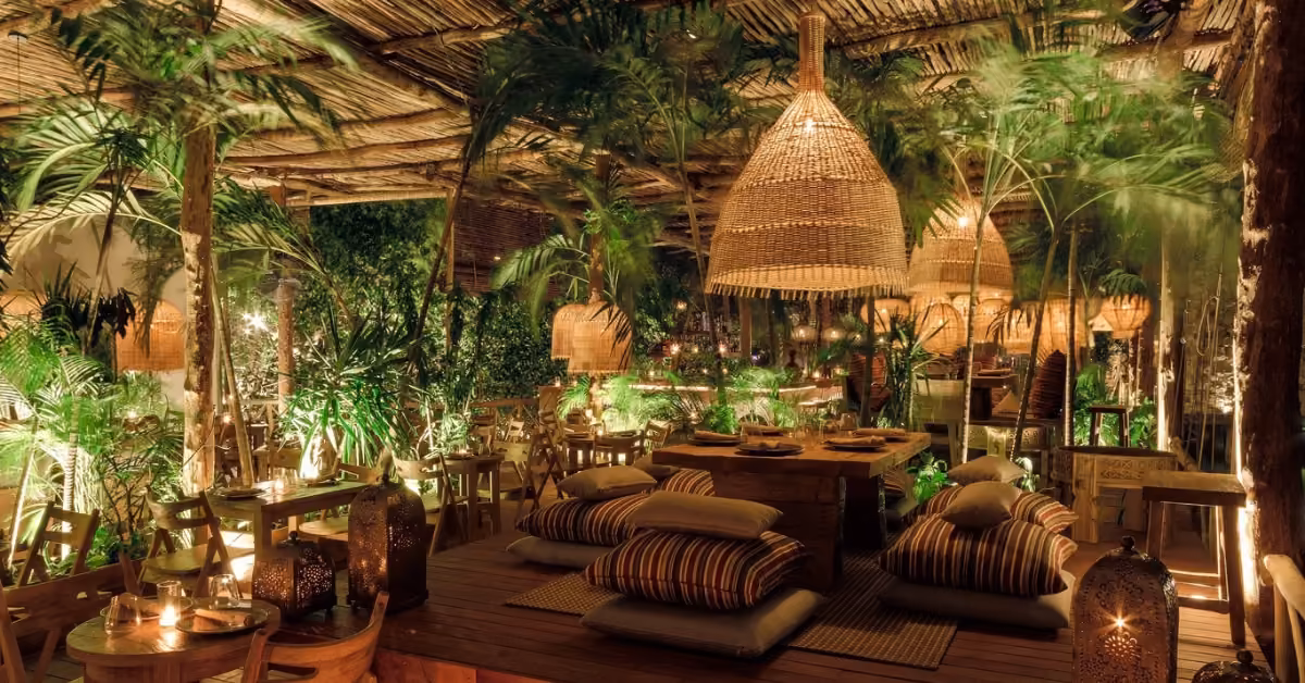 A cozy outdoor restaurant with wooden furniture, striped cushions, woven lanterns, and lush greenery under warm ambient lighting.