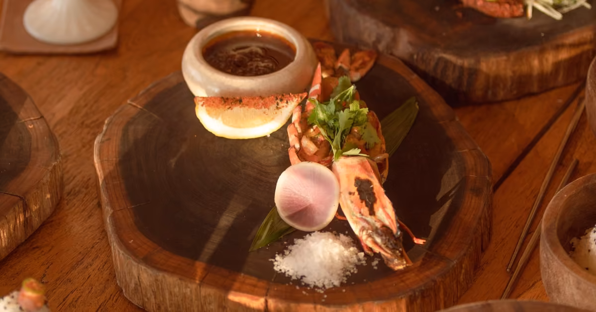 Shrimp with herbs and radish on a wooden plate, salt on the side, and dipping sauce in a stone bowl, artfully presented.