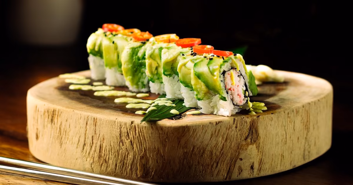 Sushi roll topped with avocado and red chili, garnished with black sesame, on a wooden board with green sauce and chopsticks.