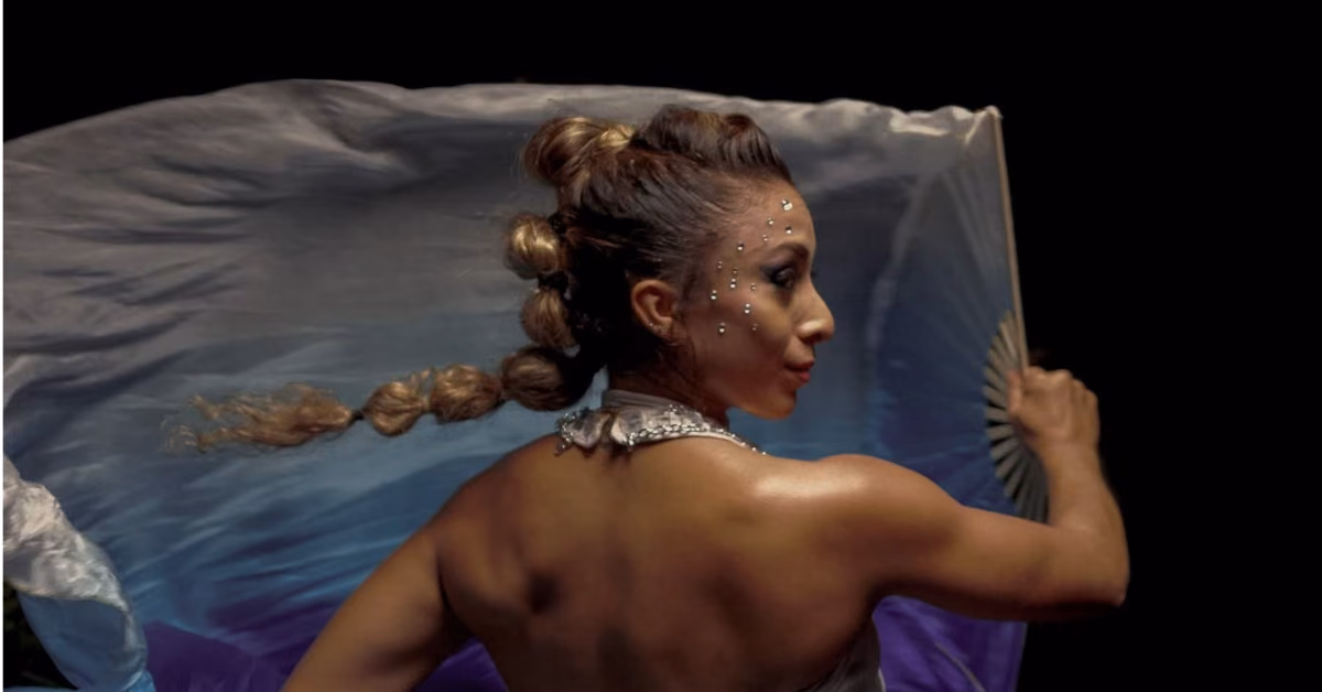 A woman with face gems and braided hair performs with a blue and white fan against a dark background, glowing with vibrant energy.