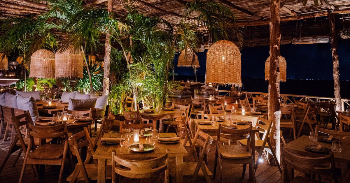 A cozy, tropical restaurant at night with wooden tables, lush plants, rattan lights, and soft candlelight for a warm ambiance.