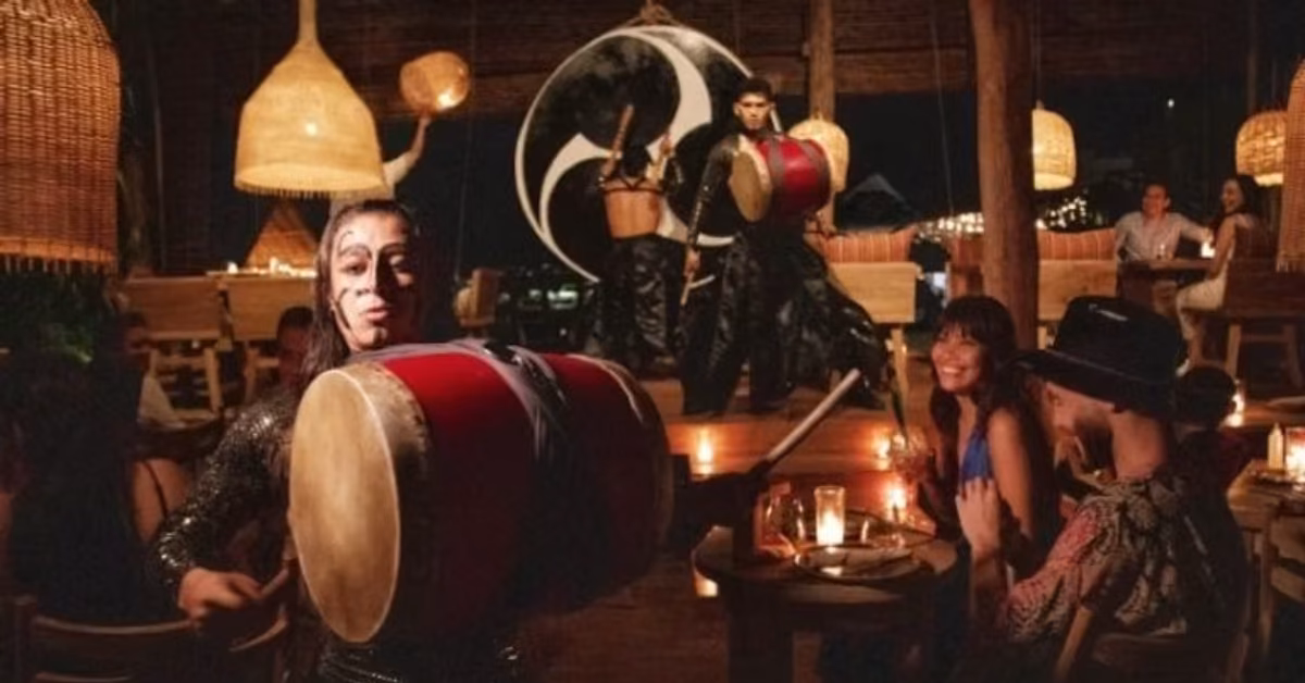 A lively restaurant with a drummer performing near diners, two drummers on stage, warm lighting, and hanging lamps.
