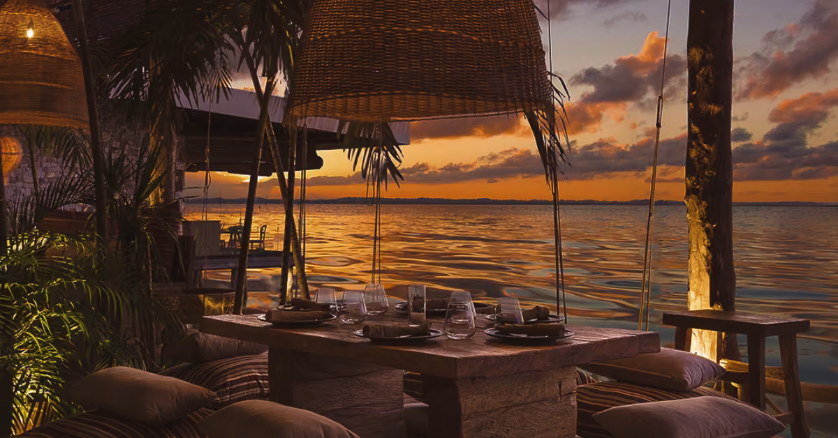 An inviting outdoor table for four with cushions, wicker lamps, and glassware overlooks a tranquil lake at colorful sunset.
